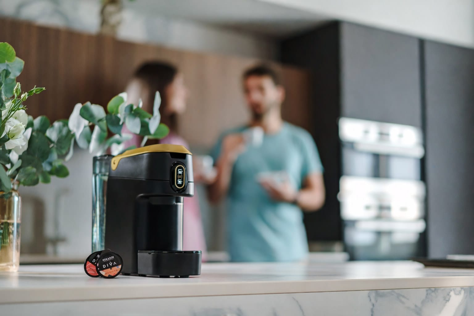 Espressor Diva Mokador cu capsule, în bucătărie; în fundal, două persoane beau cu plăcere o cafea Diva Mokador.